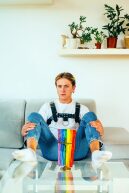 Person des LGBT-Spektrum sitzt breitbeinig da, vor ihr ein Kreuz und eine Regenbogenfahne