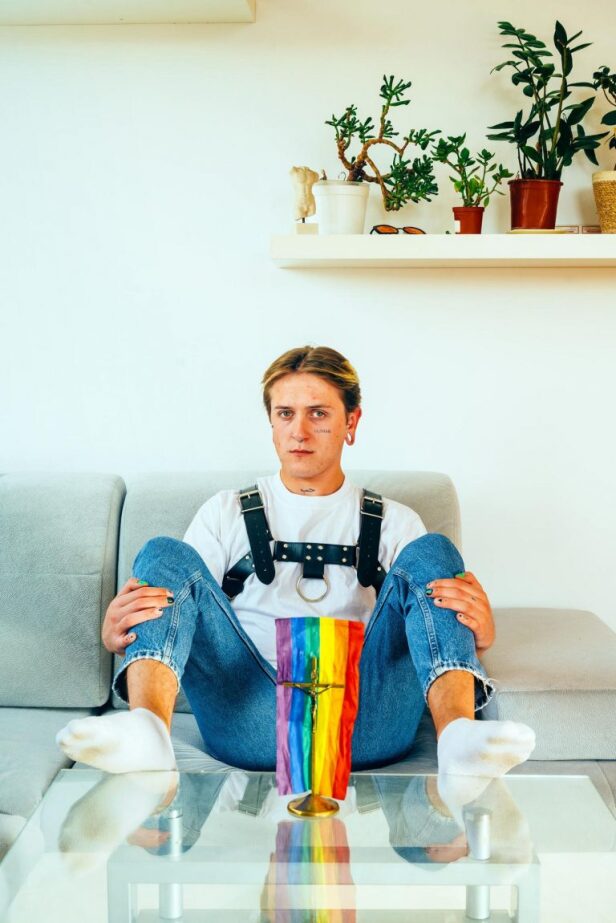 Person des LGBT-Spektrum sitzt breitbeinig da, vor ihr ein Kreuz und eine Regenbogenfahne