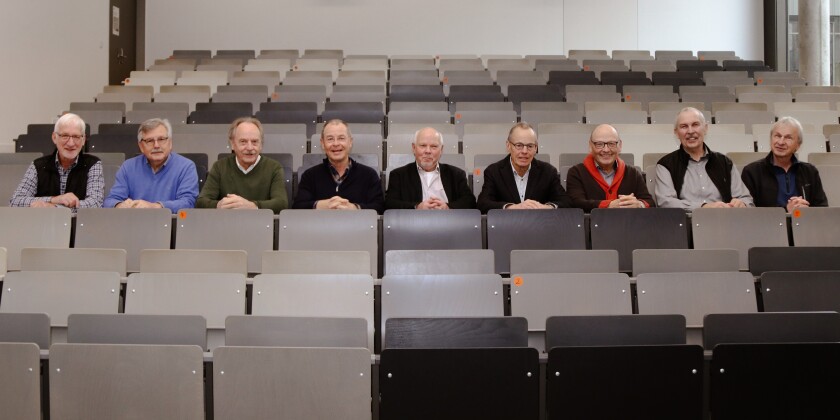 Neun Männer sitzen in einer Reihe im Hörsaal. Ansonsten ist der Hörsaal leer.