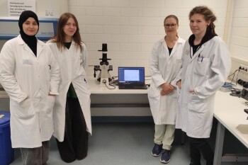 Aud dem Foto sind vier weibliche Personen in weißen Laborkitteln abgebildet. Zwei Schülerinnen stehen links, in der Mitte befindet sich ein Mikroskop, und die Labormitarbeiterinnen stehen rechts davon.