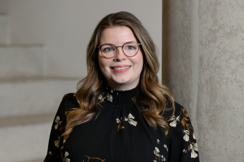 Bild von Saskia Henning die in einem schwarzen Oberteil mit Blumenmuster an eine graue Säule geleht steht. Sie trägt eine Brille und hat längere blonde haare die über ihre Schultern reichen außerdem träft sie eine brille und lächelt direkt in die Kamera