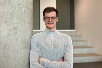 Ein Mann lehnt an einer grauen Säule. Er hat die Arme vor seinem Oberkörper verschränkt. Er hat braune Haare, eine Brille und trägt einen grauen Pullover.