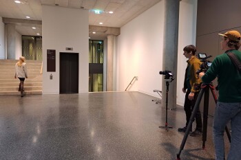 Auf einem Flur in der HSBI stehen zwei Videographen mit Kameras. Sie filmen eine Frau, die eine Treppe hochläuft.