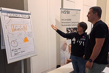 Beatriz García steht mit einem Mann vor einer Flip Chart. Sie erklärt ihm das Projekt InCamS@BI. Beatriz trägt einen schwarzen Pillover der HSBI, hat kinnlanges dunkles Haar und trägt eine Brille. Ihr Gesprächspartner trägt ein schwarzes T-Shirt.