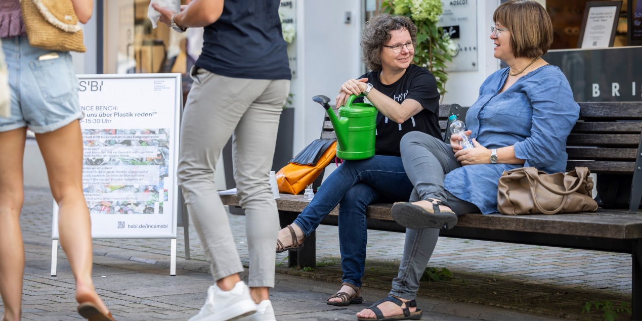 Heike Wulf sitzt in einem schwarzen T-Shirt auf einer Bank in der Bielefelder innenstadt neben ihr sitzt eine Frau mittleren Alters in einem hellblauen Jeanshemd die beiden unterhalten sich sie haben ihre beine übereinandergeschlagen und schauen sich an