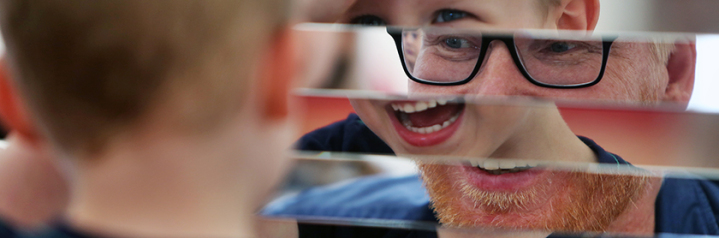 Im unscharfen Bildvordergrund sieht man am linken Bildrand den blonden Hinterkopf eines Kindes es schaut auf eine Spiegelfläche die an mehreren Stellen durchlässig ist durch diese länglichen Lücken im Spiegel guckt ein männliches Gesicht mit Brille