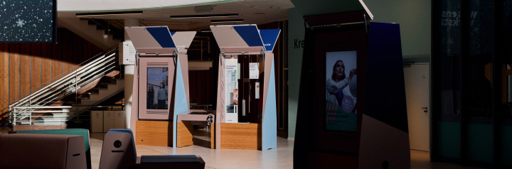 Bild vom sogenannten Marktstand der HSBI in der Wissenswerkstadt Der Stand besteht aus drei Teilen linkst steht ein schrankähnliches Gehäuse in dem ein Hochkantbildschirm angebracht ist in der Mitte befindet sich ein flacher Tisch mit einem Touchbildschir