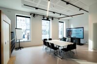 Bild des Studios im Erdgeschoss der Wissenswerkstadt in der Mitte des kleinen Raums ist ein Gruppentisch mit 10 Stühlen aufgebaut durch die großen Fenster fällt tageslicht ein im rechten Bildteil ist eine großformatige Videoleinwand zu sehen