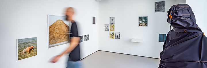Blick in den Ausstellungsraum HSBI-Satellit Vor einer weißen Wand mit Bildern läuft ein Mann in einem schwarzen T Shirt rechts von ihm seht eine Figurine mit einem Umhangähnlichen Gewand