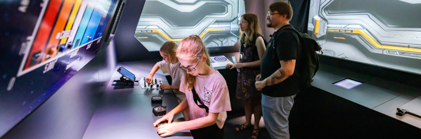 Bild einer Gruppe von vier Personen die in der Wissenswerkstadt das Escape Game spielen im Vordergrund sieht man einen Jungen und ein Mädchen im Alter von circa 7 Jahren an einem verschiedenen Knöpfen und Hebeln spielen über ihnen und im hintergrund des b