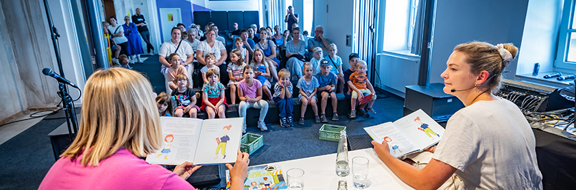 Bild einer Lesung in der Wissenswerkstadt: an einem Tisch im Bildvordergrund sitzen zwei Frauen mit dem Rücken zur Kamera im Bildhintergrund sieht man mehrere Stuhlreihen auf denen das Publikum der Lesung sitzt darunter viele Kinder