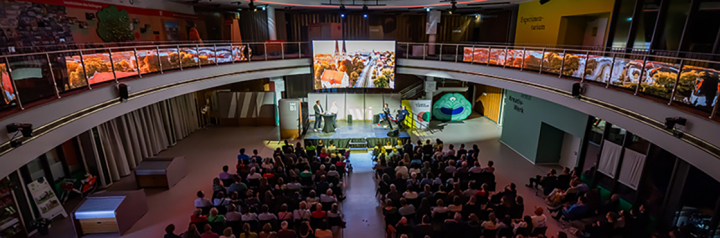 Bild des zentralen Raums im Erdgeschoss der Wissenswerkstadt genannt Oval bei einer Abendveranstaltung der Raum ist abgedunkelt im bildhintergrund sieht man eine hell erleuchtete Bühne vor der im bildvordergrund zuschauer innen in mehreren reihen sitzen