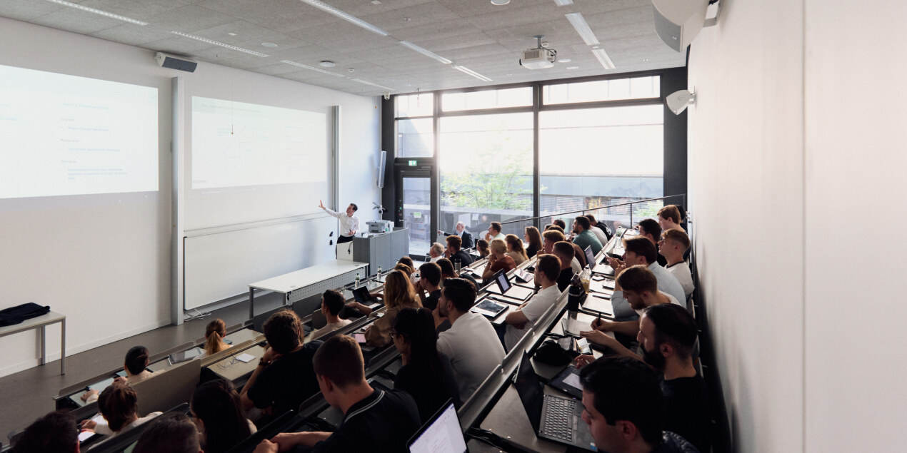 Foto: Ein vollbesetzter Hörsaal an der Tafel erklärt ein Dozent etwas