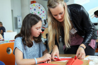 Frau zeigt Mädchen wie man einen Papierflieger bastelt.