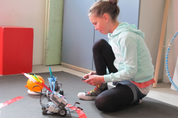 Antonia Flößer hockt am Boden mit dem Roboter vor sich und einem Smartphone zur Steuerung in ihrer Hand.