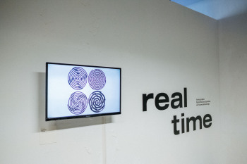 Auf einer weißen Wand steht der Schriftzug Real Time daneben hängt ein Bildschirm auf dem sich vier unterschiedlich gemusterte Kreise befinden