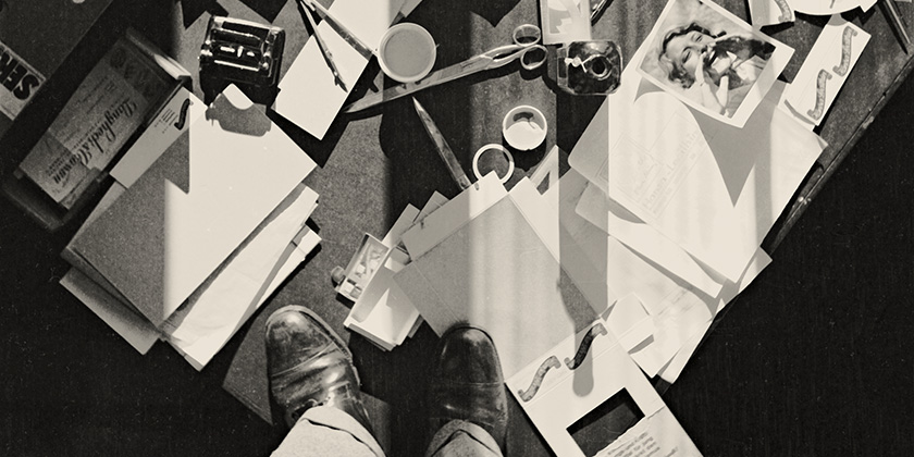 Auf einem Schreibtisch liegen verschiedene Büromaterialen chaotisch umher. Auf dem Schreibtisch steht ein Mann.