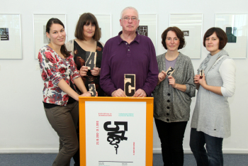 Professor Jochen Geilen steht mit vier Studierenden an einem Rednerpult. Sie halten Druckplatten in den Händen,auf denen Zahlen zu sehen sind. Im Hintergrund sind Druckgrafiken der Ausstellung zu sehen.