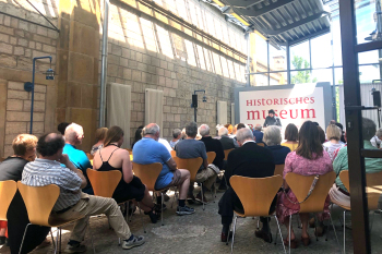 Auf Stuhlreihen sitzen Menschen und schauen auf einen Mann der vor einer Wand mit dem Schrifzug Historisches Museum steht