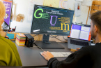 Menschen sitzen vor einem Bildschirm, darauf steht in großen bunten Buchstaben GUM