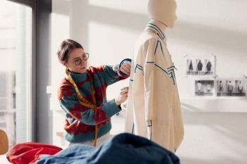 Eine Frau steht an einer Puppe und klebt ein Kleidungsstück mit Klebeband ab.