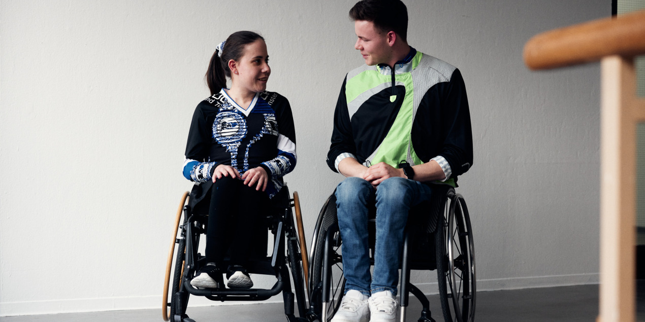Eine Frau und ein Mann im Rollstuhl tragen die Modeentwürfe der Studierenden