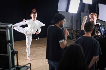 Eine Tänzerin in weißem Kostüm mit Schulterpolstern tanzt im Scheinwerferlicht Im Vordergrund ist Kameraequipment zu sehen