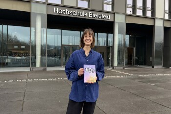 Carina Thomas steht mit ihrem Buch vor der Hochschule Bielefeld und lächelt