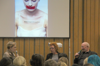 Dr. Anna Zika, Prof. Willemina Hoenderken und Karl Martin Holzhäuser diskutieren auf einem Podest in der Alten Stadtbibliothek miteinander. Im Hintergrund ist eien Projektion zu sehen, in der eine Frau ein weißes Kleid trägt und über dem Mund einen roten