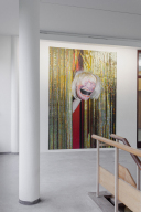 An einer Wand hängt eine große Fotografie, auf der eine Hadn eine grostesk lachende Maske durch einen goldenen Vorhang streckt