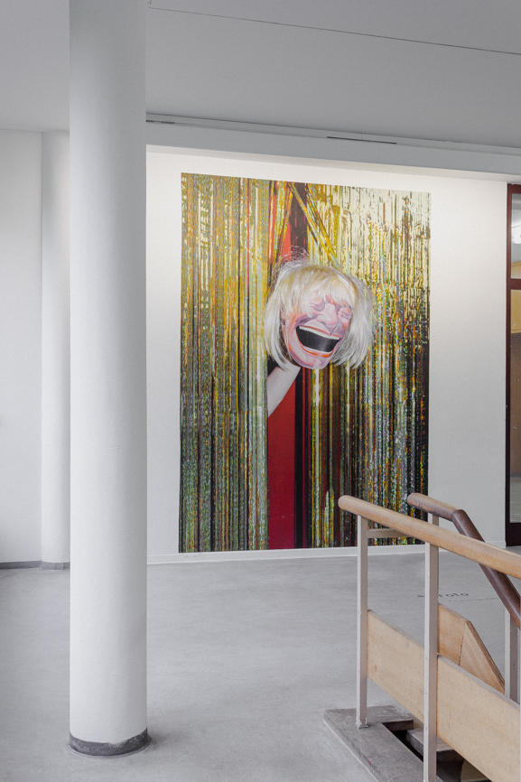 An einer Wand hängt eine große Fotografie, auf der eine Hadn eine grostesk lachende Maske durch einen goldenen Vorhang streckt