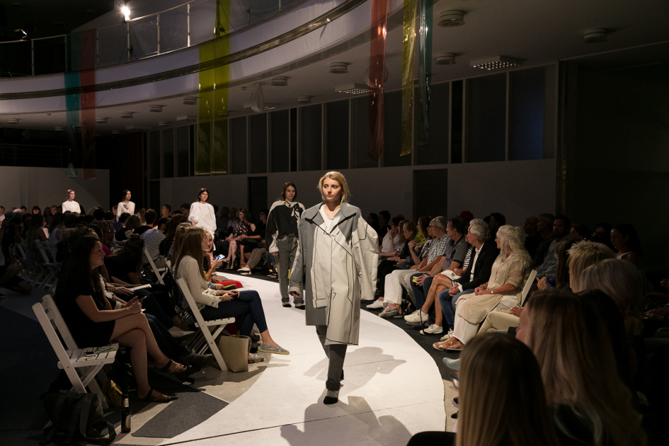 Personen gehen über einen Laufsteg. Am Rand sitzt das Publikum auf Stühlen.