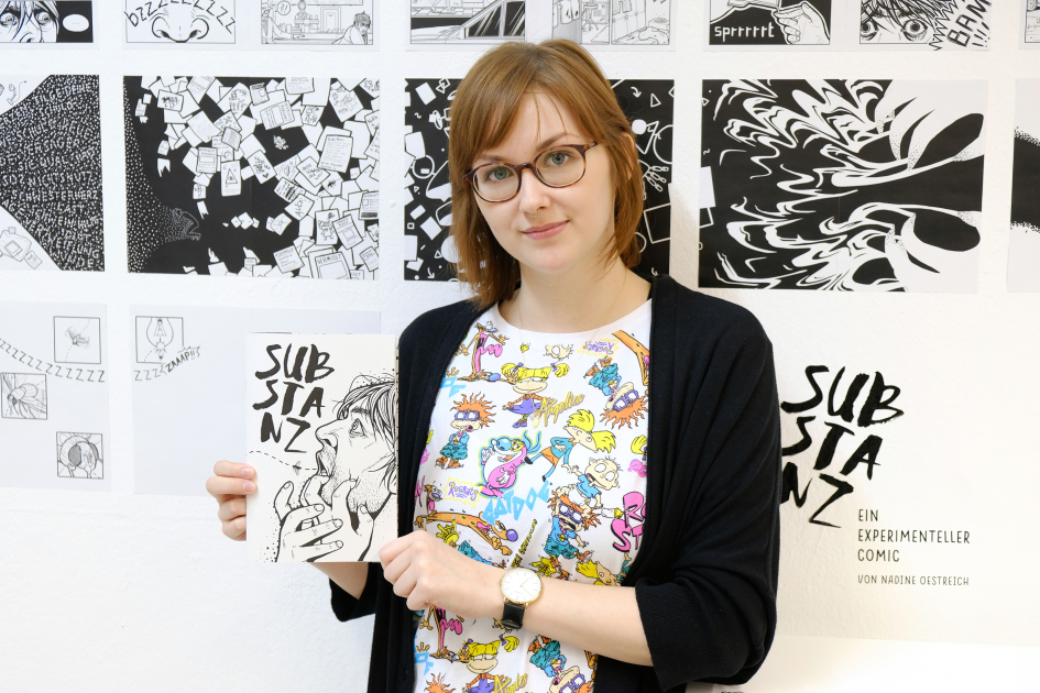 Frau hält Comic in den Händen und steht vor einer Wand mit einzelnen Comic-Seiten.