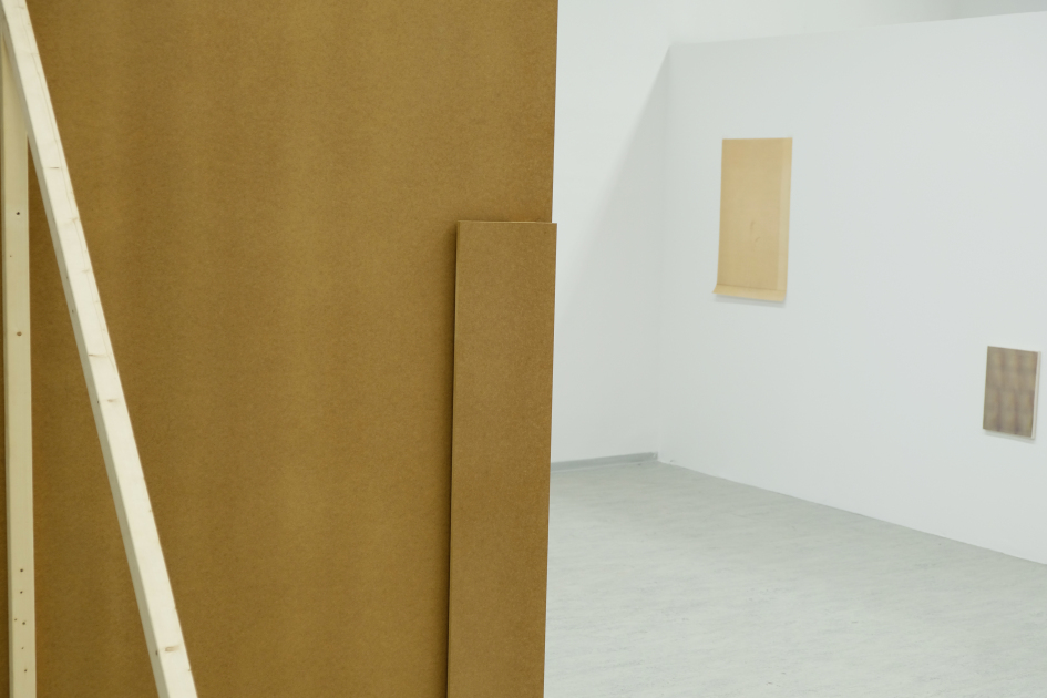 Im Vordergrund eine Holzwand, im Hintergrund an der Wand hängen Bilder.