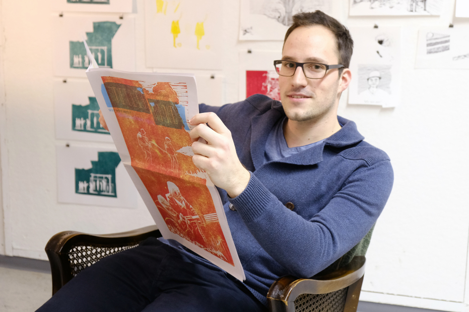 Männliche Person sitzt auf einem Sessel und hält eine Art Zeitung in seinen Händen.