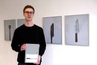 Ein Mann hält ein Buch und steht vor einer Wand mit Bildern von Messern.