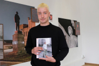 Eine Person hält ein Buch und blickt in die Kamera.