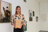 Eine Person hält ein Buch und blickt in die Kamera.