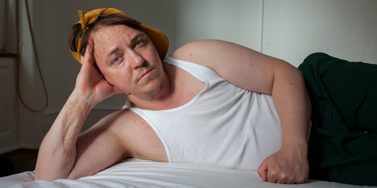 Eine Person liegt in Unterhemd auf einem Bett und schaut in die Kamera