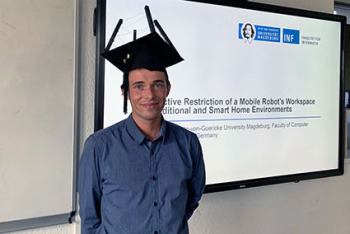 Ein Mann mit Doktorhut steht vor einem Bildschirm und lächelt in die Kamera.