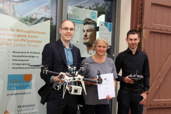 Zwei Studenten mit der Professorin in der Mitte stehen vor einem Gebäude und halten die Helikopterdrohne und die Fernsteuerung in der Hand.