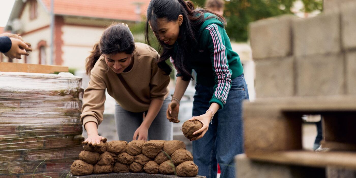 zwei Studentinnen formen Lehmkugeln und schcihten sie zu einer kleinen Mauer