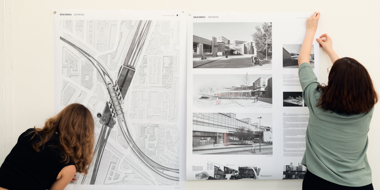 Zwei Studentinnen hängen ein Plakat mit Entwürfen an eine Wand