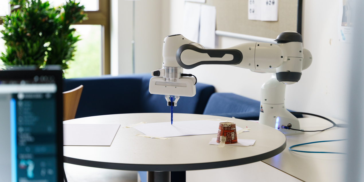 Ein Roboterarm zeichnet mt einem Stift etwas auf ein Blatt Papier
