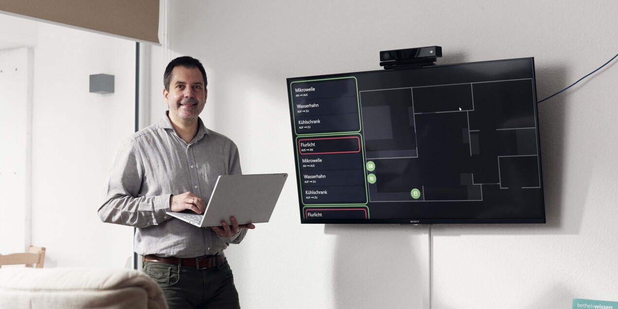 Prof. Dr. Thorsten Jungeblut steht neben einem Bildschirm, auf dem ein Grundriss der Kogni-Home Wohnung zu sehen ist.
