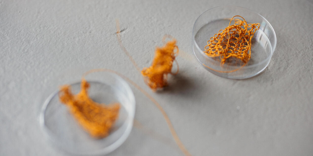 Detailaufnahme von drei kleinen gehäkelten Textilien die auf einem grauen Untergrund liegen Die Häkeltextilien bestehen aus orangenem Garn und haben die Formen eines T sowie einer Röhre