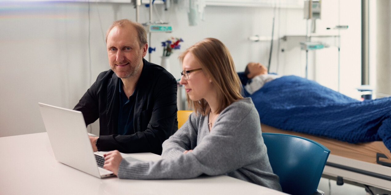 Professor Doktor Horst schaut in die Kamera während er mit Chiara Freitag am Rechner sitzt
