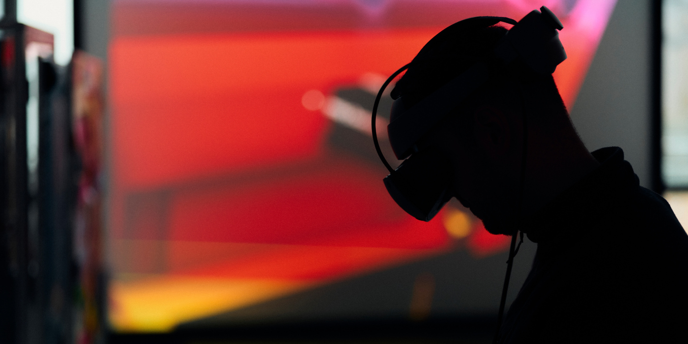 Vor einem mehrfarigen Hintergrund der von unten nach oben die Farben gelb rot pink abbildet sieht man den Schattenumriss eines Oberkörpers Auf dem Kopf trägt die Person eine Virtual Reality Brille Sie hat den Kopf leicht nach unten geneigt