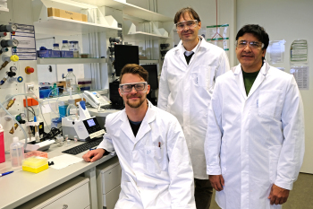 Ein Mann in einem weißen Laborkittel sitzt auf einem Stuhl in einem Labor. Hinter ihm stehen zwei Männer ebenfalls in Laborkitteln