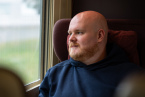 Porträtbild von Johannes Reinholz der Student des Studiengangs Digitale Bahnsysteme steht in einem blauen Kapuzenpullover sitzend in einem ausrangierten ICE Zug er hat eine Glatze und einen roten Vollbart und schaut aus dem Fenster